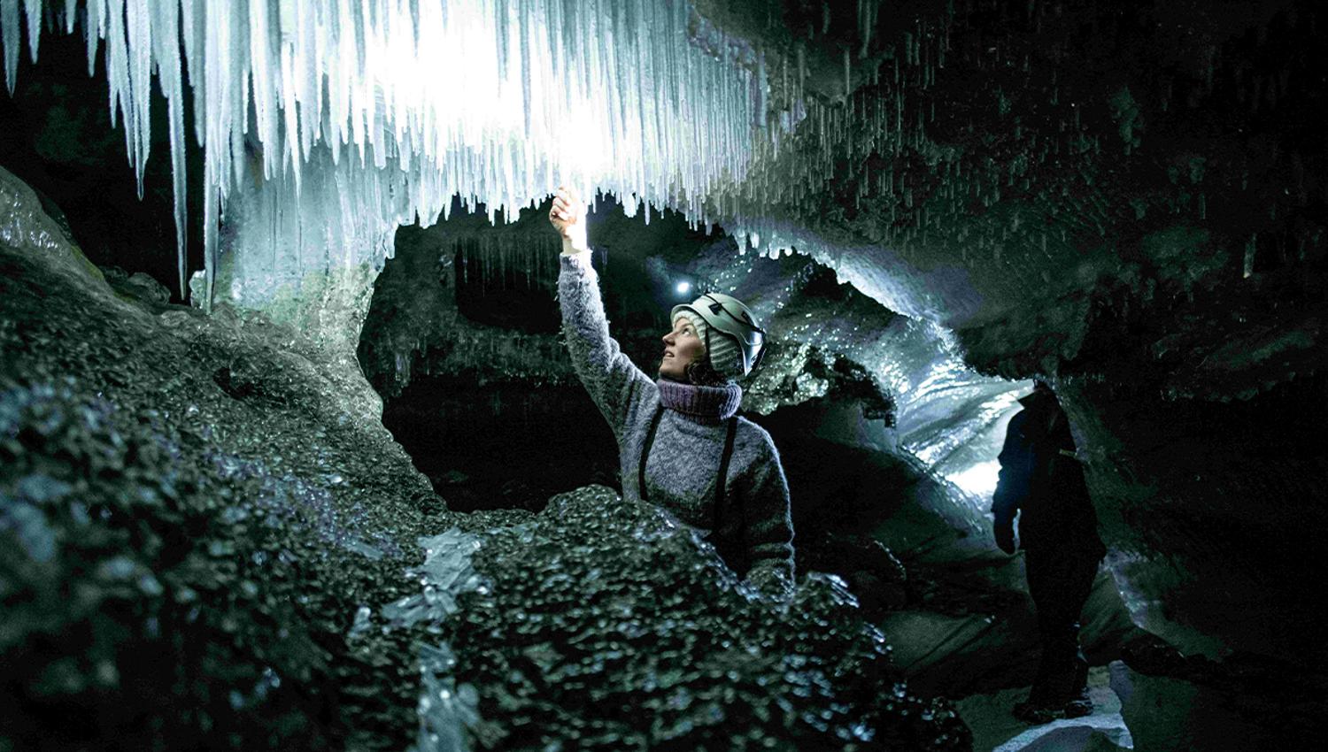 A guide with a bright head lamp and helmet inside an ice cave feeling one of the icicles hanging from the cave roof
