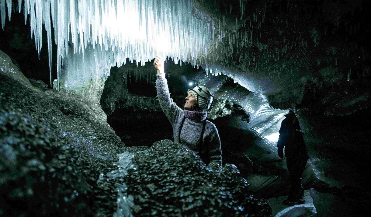 A guide with a bright head lamp and helmet inside an ice cave feeling one of the icicles hanging from the cave roof