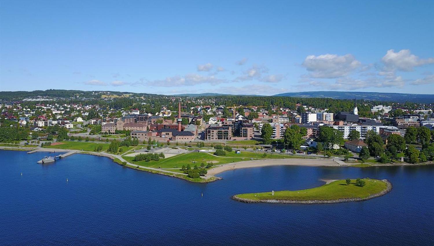 Oversiktsbilde av en by ved et stort vann, med grønt landskap og spredte skyer på himmelen.
