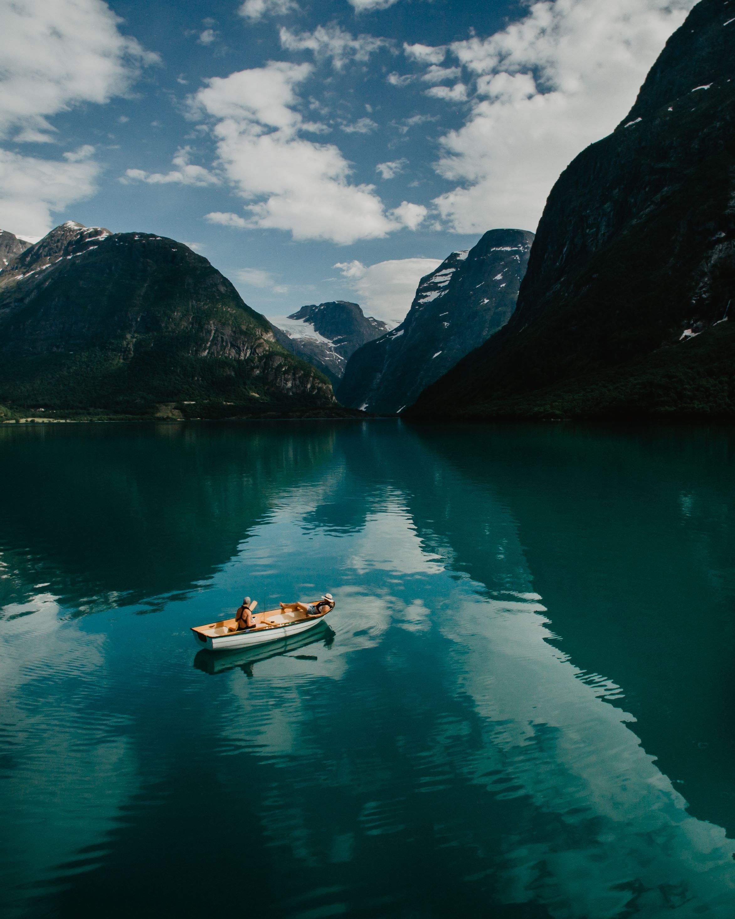 Ei lita robåt flyt på turkis fjordvatn mellom bratte fjell, med skyer og speglingar i overflata..