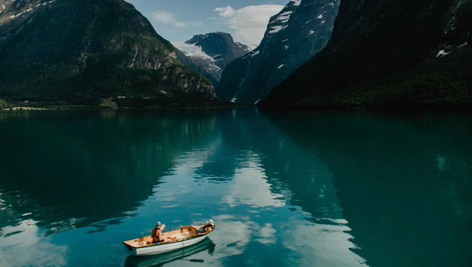 Ei lita robåt flyt på turkis fjordvatn mellom bratte fjell, med skyer og speglingar i overflata..
