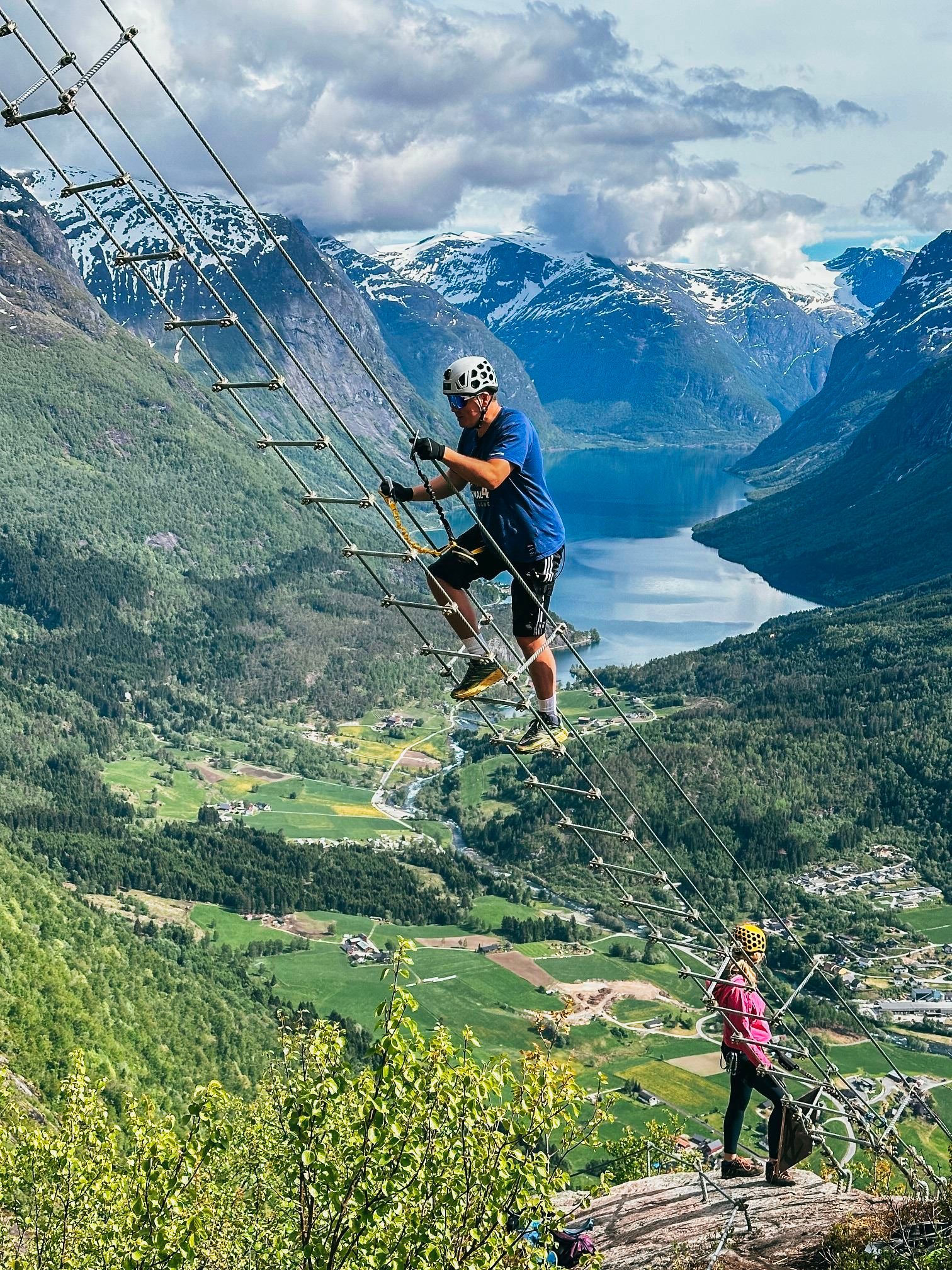 To klatrarar i hjelm på ein via ferrata-stige langs fjellveggen med fjord, dal og snødekte fjell i bakgrunnen..