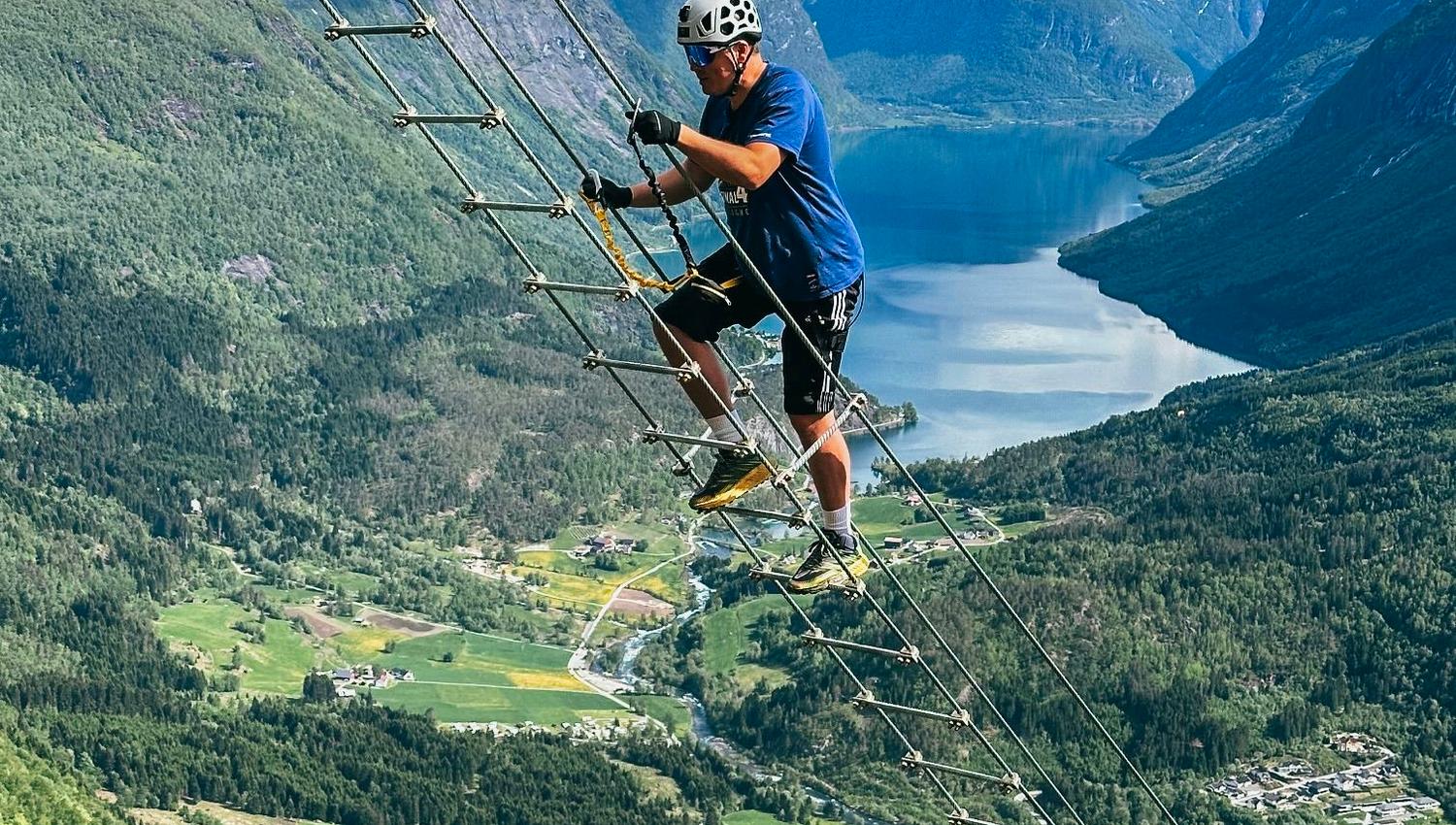 To klatrarar i hjelm på ein via ferrata-stige langs fjellveggen med fjord, dal og snødekte fjell i bakgrunnen..
