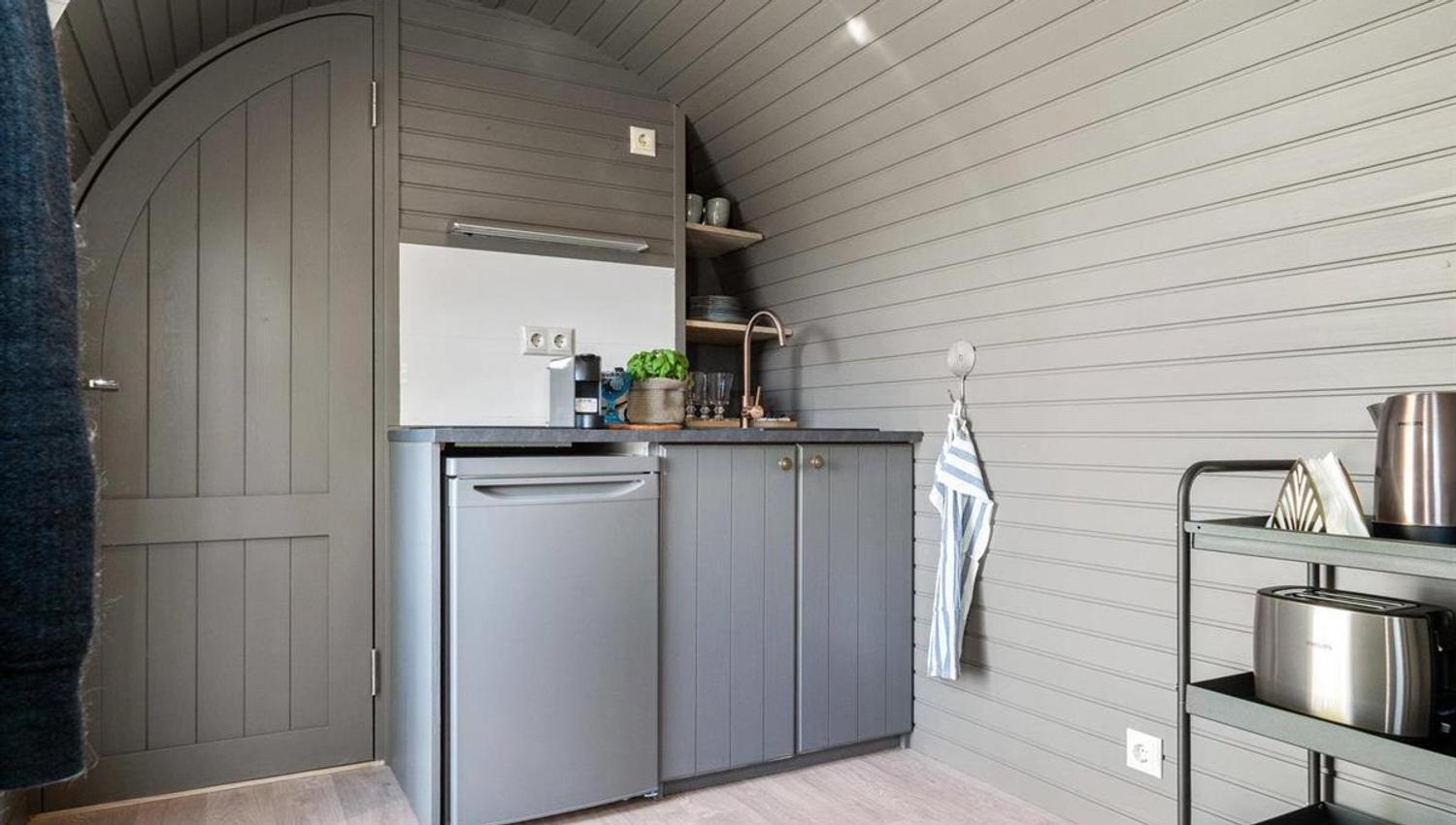Small kitchenette with a fridge and storage inside a wooden cabin pod.