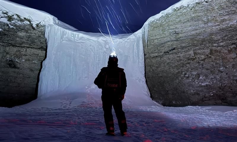 A person is standing in the dark with a headlamp, ligting up in the sky towards a frozen waterfall with snowy weather