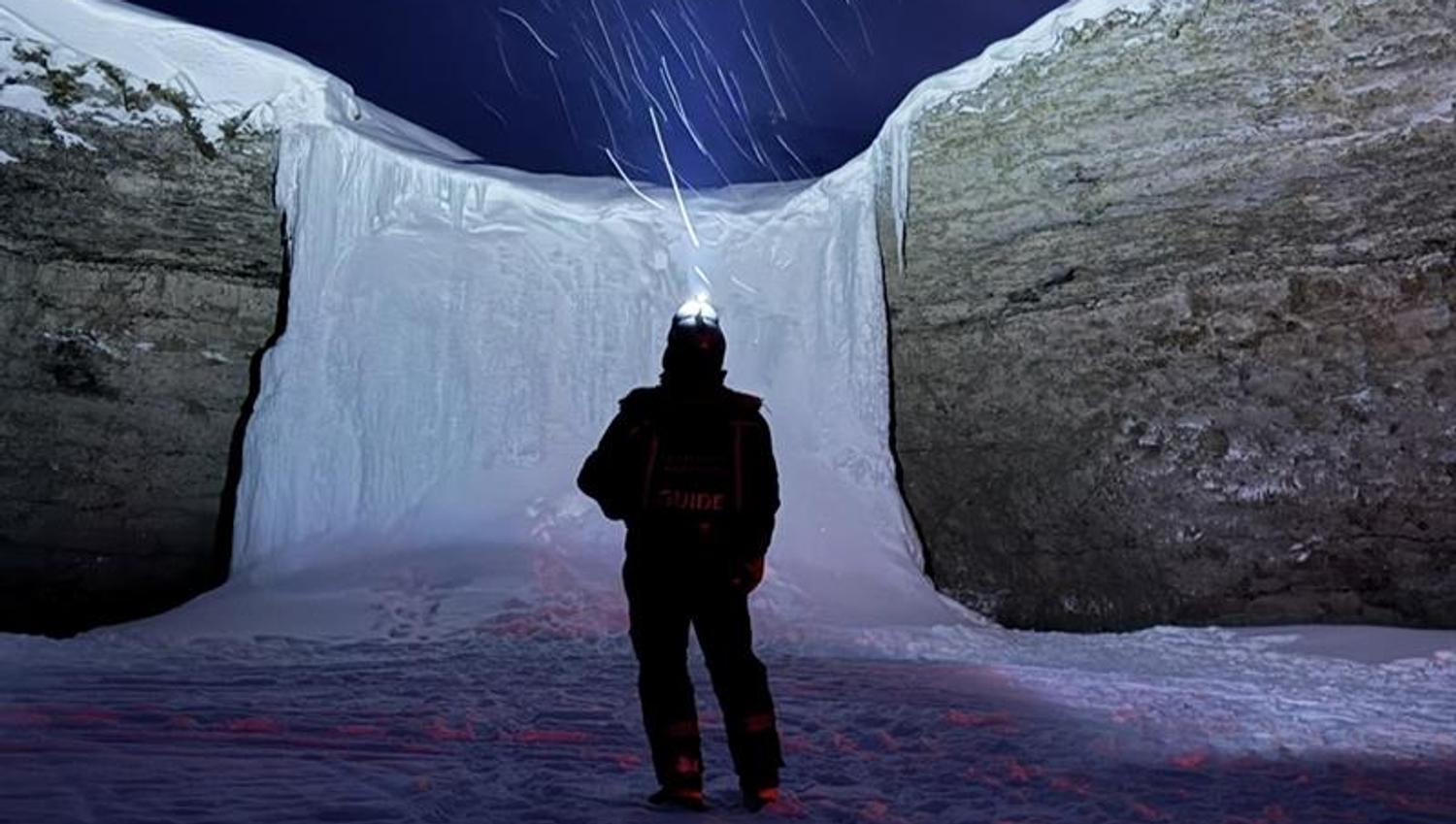 A person is standing in the dark with a headlamp, ligting up in the sky towards a frozen waterfall with snowy weather