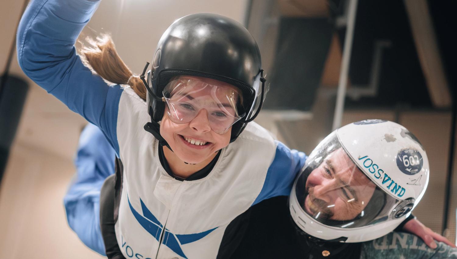 Young flyer in a VossVind jumpsuit and helmet, arms outstretched mid‑flight in the vertical wind tunnel, supported by an instructor.