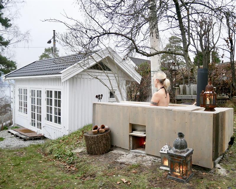 Lite, hvitt tiny house med sovehems og store vinduer, plassert i en grønn hage med trær. person i et badekar foran huset med en terrasse med utsikt mot fjorden.