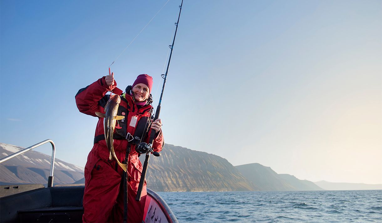 A guest standing in a RIB boat holding a fishing pole and a caught fish, with an open fjord and mountains in the background