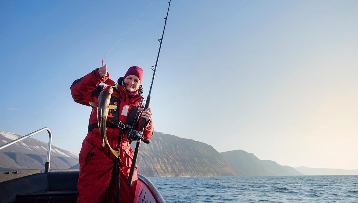 A guest standing in a RIB boat holding a fishing pole and a caught fish, with an open fjord and mountains in the background