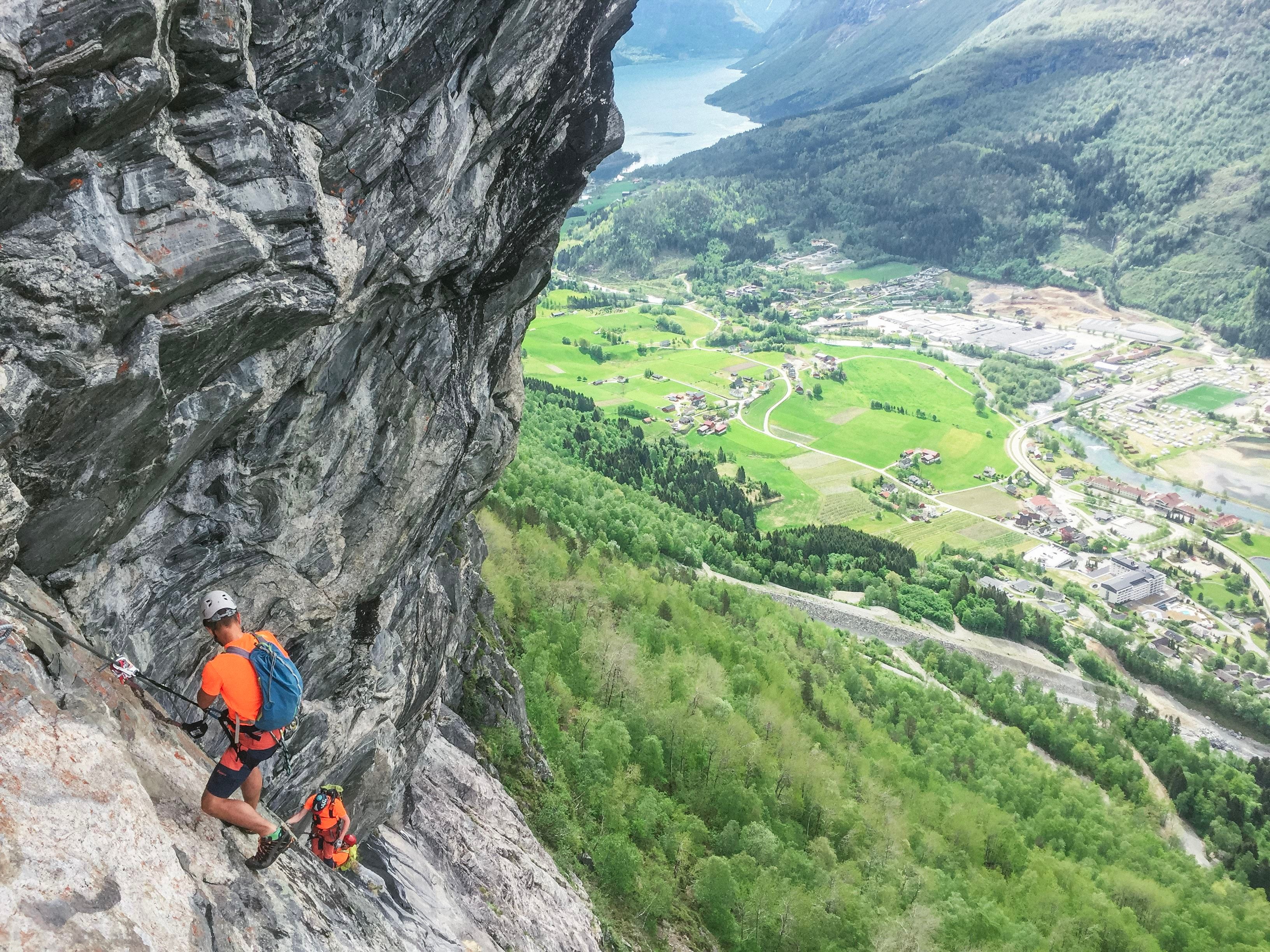 To klatrarar med hjelm og ryggsekk på ei bratt fjellside, med grøne dalar, gardar og fjord i bakgrunnen..
