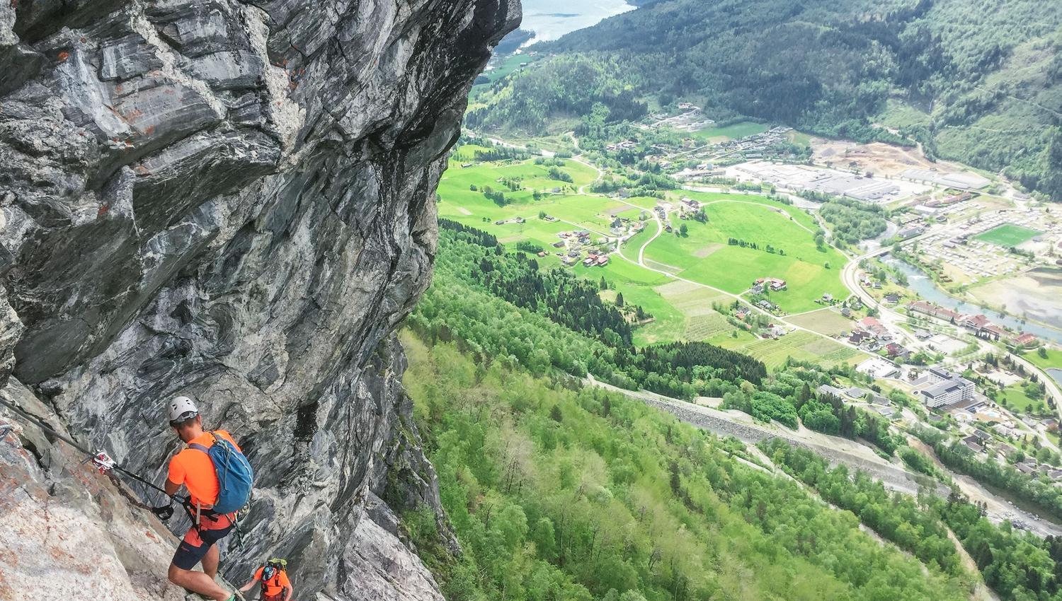 To klatrarar med hjelm og ryggsekk på ei bratt fjellside, med grøne dalar, gardar og fjord i bakgrunnen..