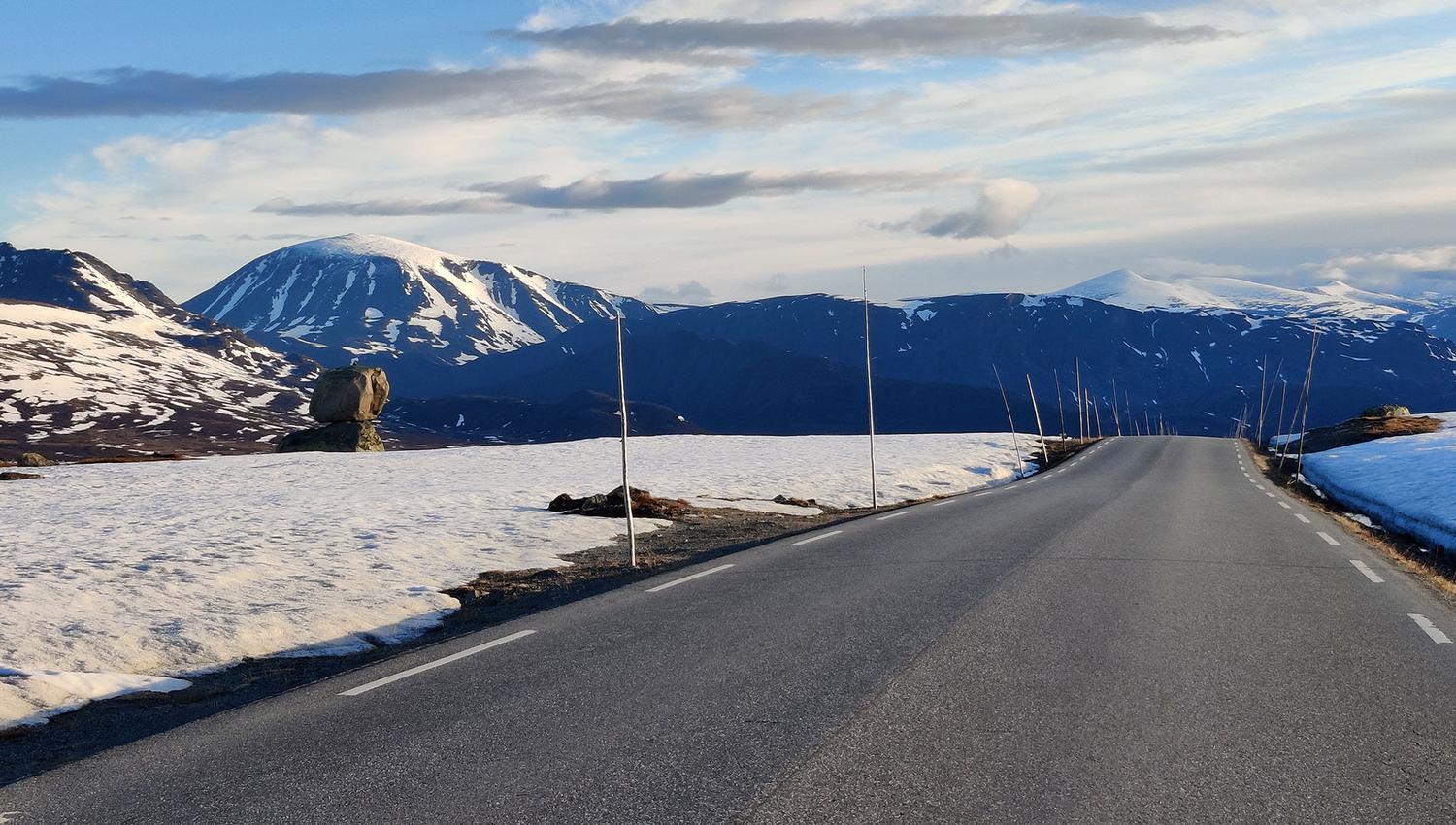 Vei over snødekt fjellplatå med snødekte fjell i bakgrunnen.