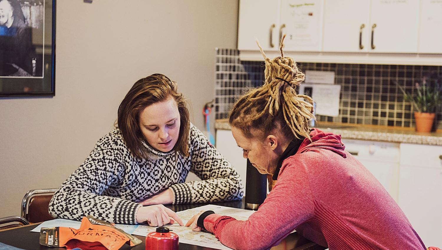 Two guests sitting at a table in a shared kitchen area at Coal Miners' Cabins planning a trip, with various outdoors equipment laid out on the table