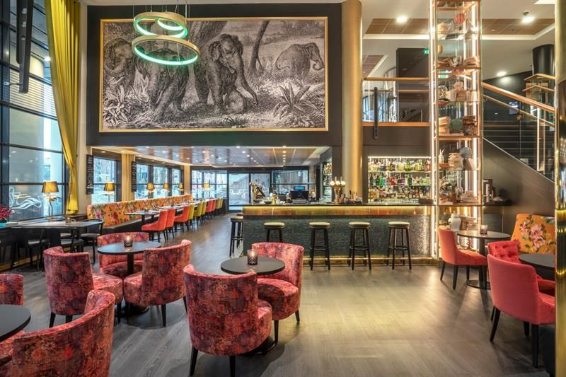 A bar with bar stools, small round tables with red chairs, a large mural of elephants hanging on the wall, and a staircase in the background