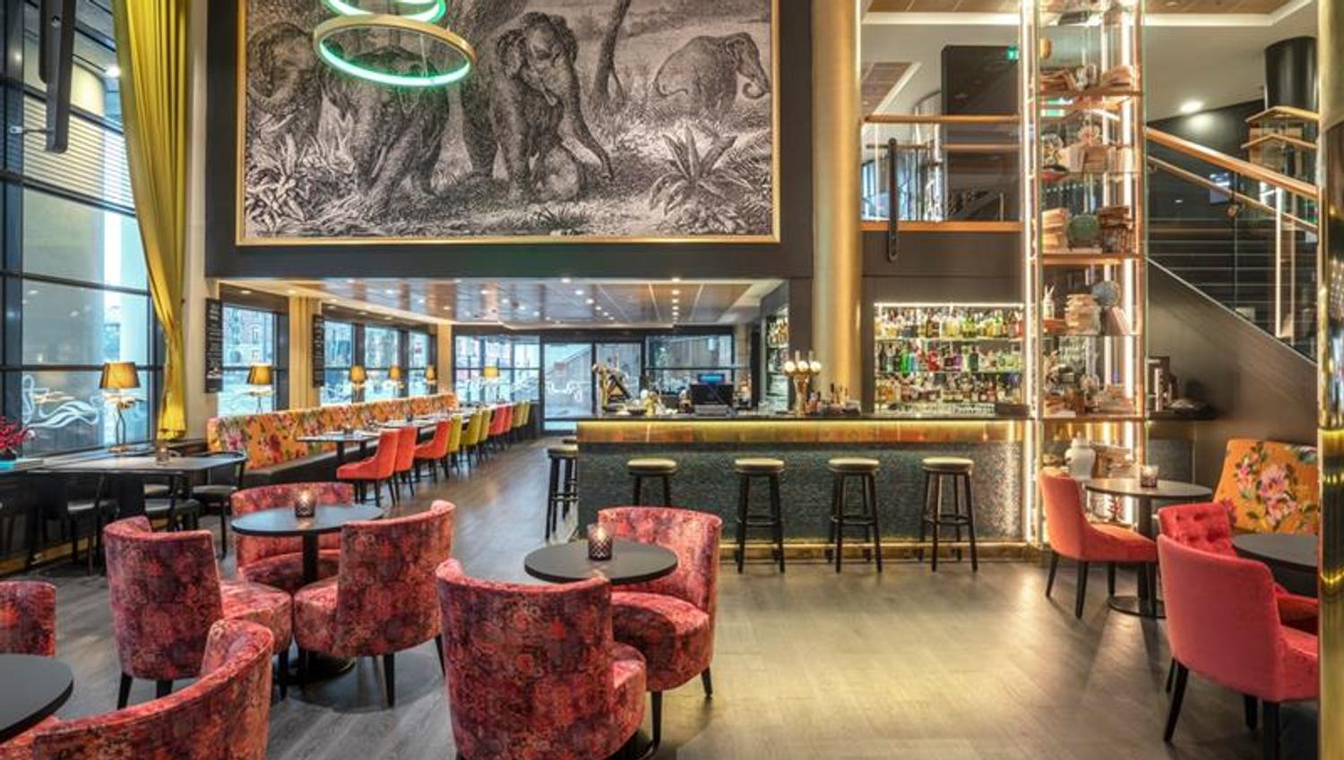 Bar med barstoler, små runde bord med røde stoler, et stort veggmaleri av elefanter som henger over og trapp i bakgrunnen