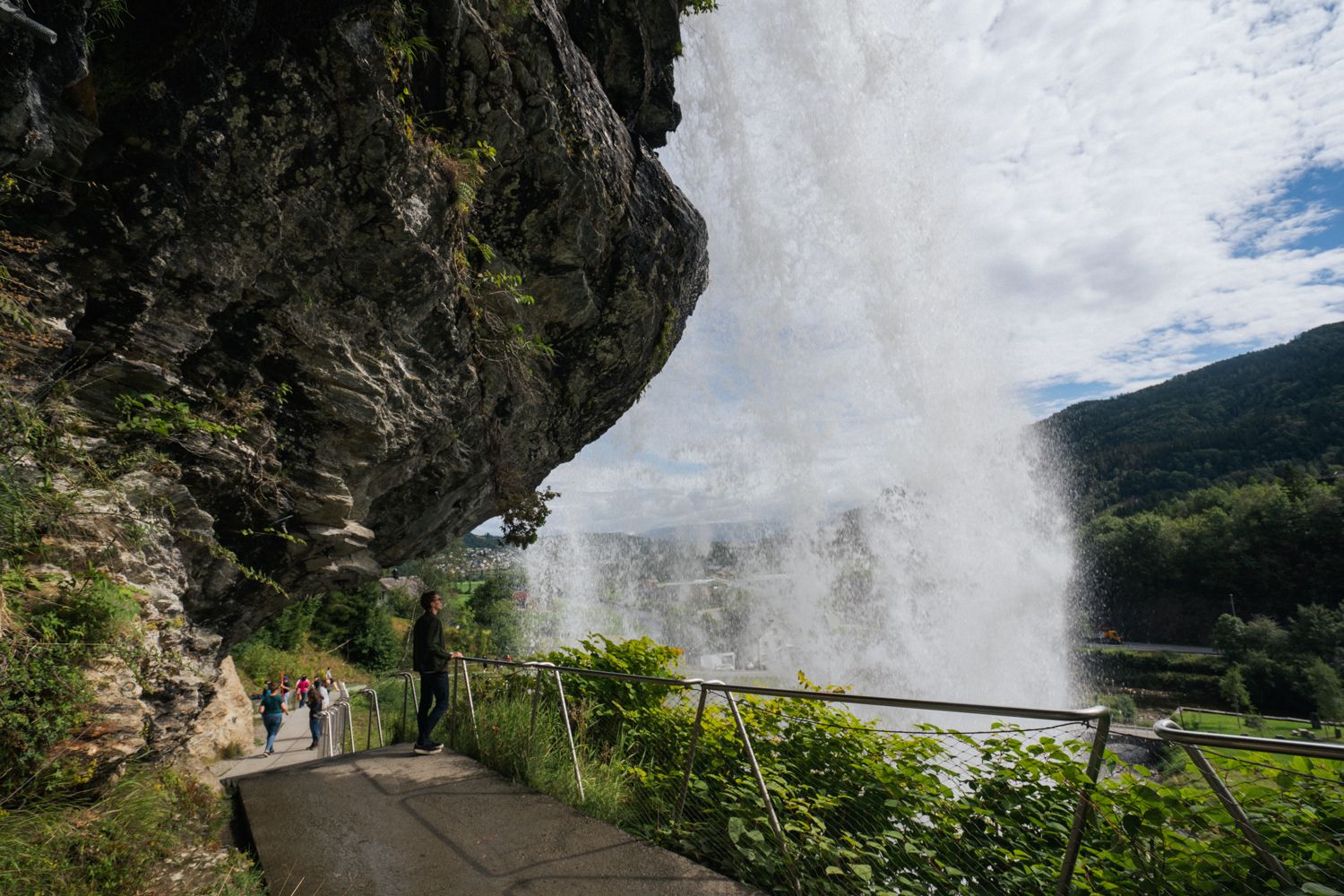 Nærbilde av besøkende som går under Steinsdalsfossen på en tilrettelagt sti, med vannet som faller kraftig i bakgrunnen og den norske naturen i full visning.