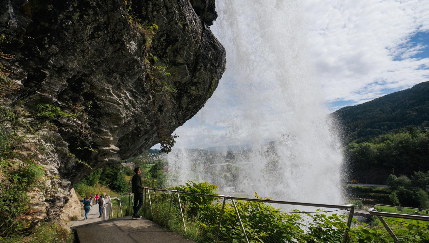 Nærbilde av besøkende som går under Steinsdalsfossen på en tilrettelagt sti, med vannet som faller kraftig i bakgrunnen og den norske naturen i full visning.