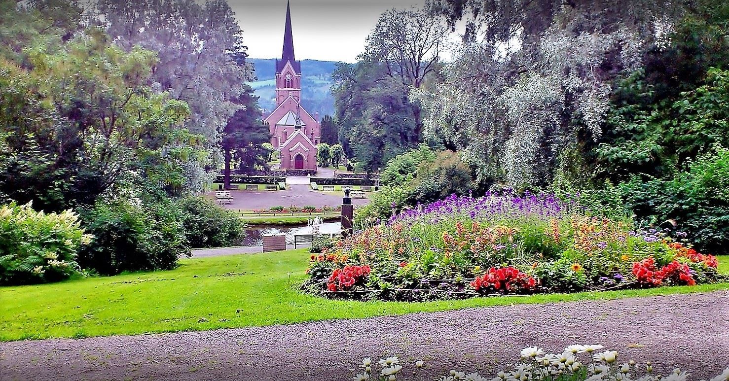 Utsikt gjennom en park med blomsterbed og trær mot en rød kirke med høyt spir, med åser i bakgrunnen..