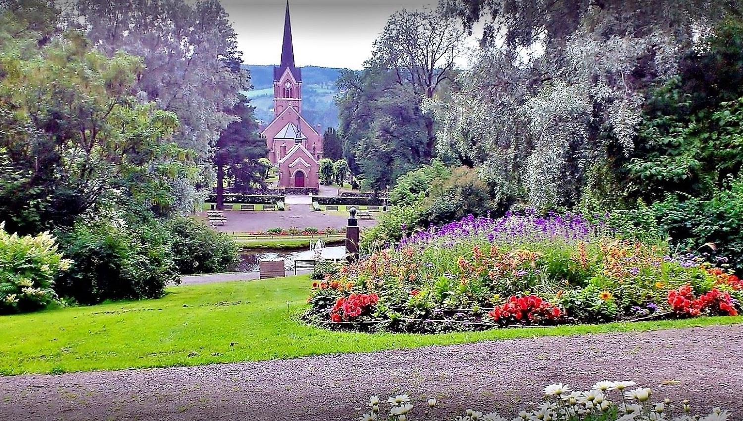 Utsikt gjennom en park med blomsterbed og trær mot en rød kirke med høyt spir, med åser i bakgrunnen..