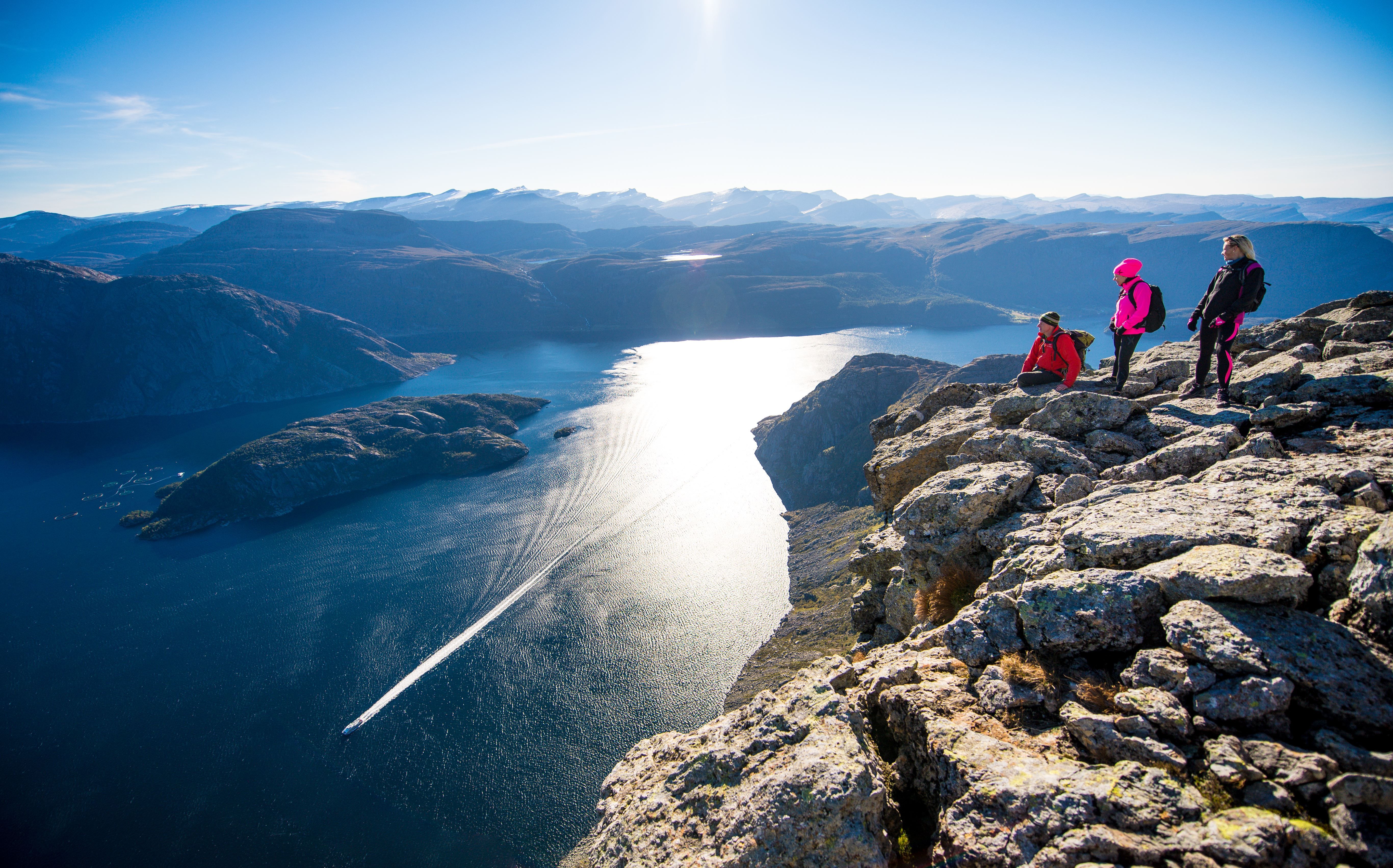 Three hikers stand on a rocky cliff overlooking a sunlit fjord with a boat wake, surrounded by distant mountains..