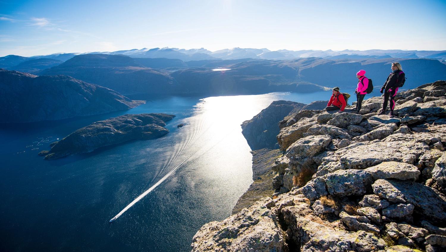 Three hikers stand on a rocky cliff overlooking a sunlit fjord with a boat wake, surrounded by distant mountains..