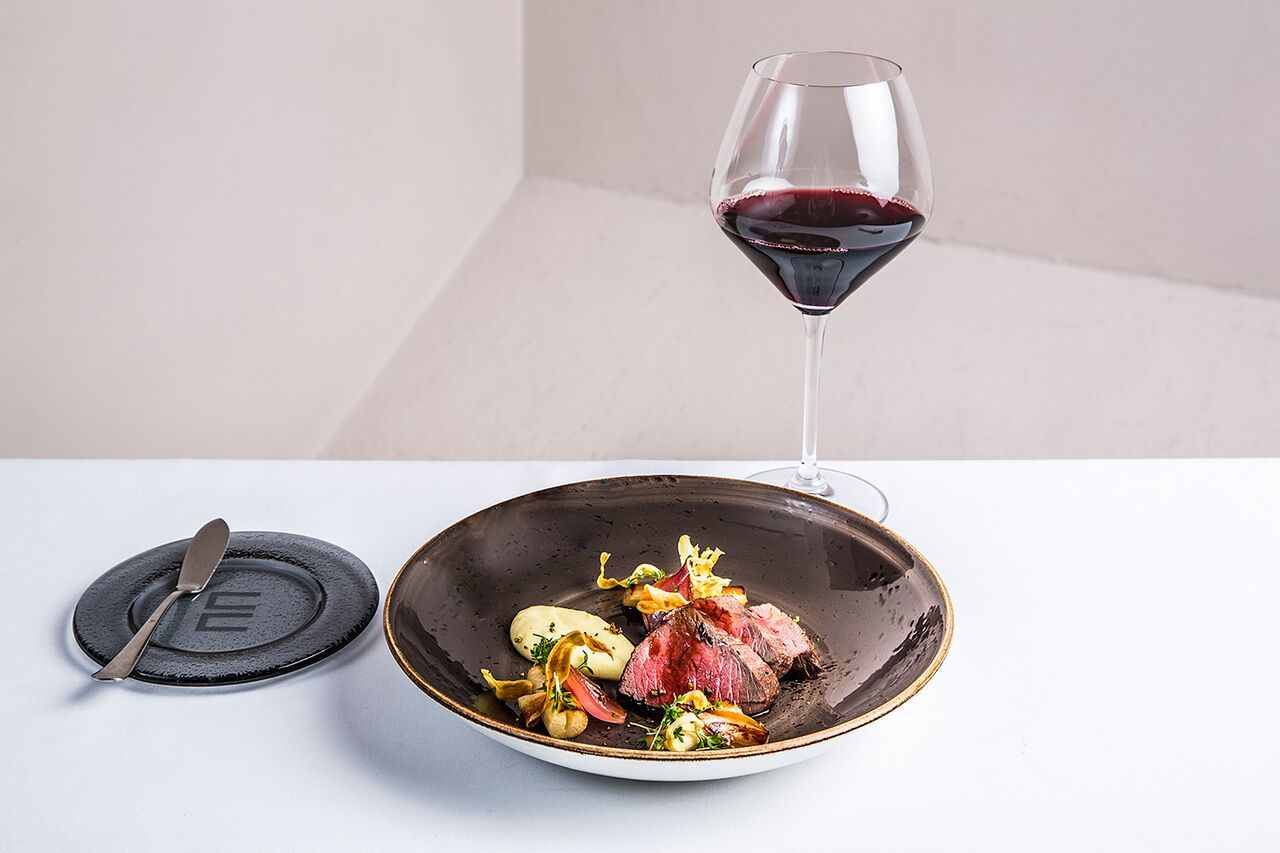 Detail photo of a dish in a brown bowl, next to a glass of red wine. White background.