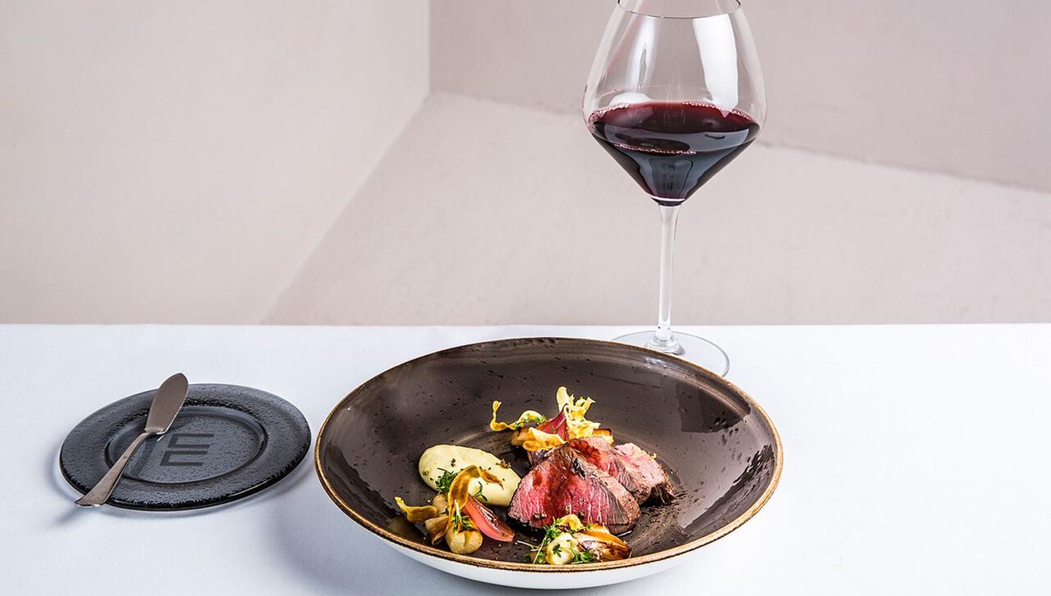 Detail photo of a dish in a brown bowl, next to a glass of red wine. White background.