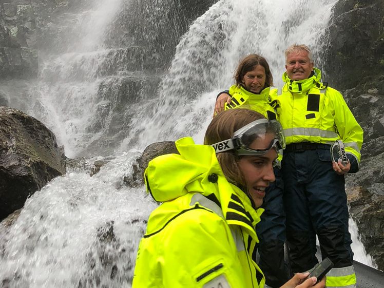 Spente deltakarar står tett inntil fossen og kjenner den kraftige vasspruten på RIB-safari i Hardanger.