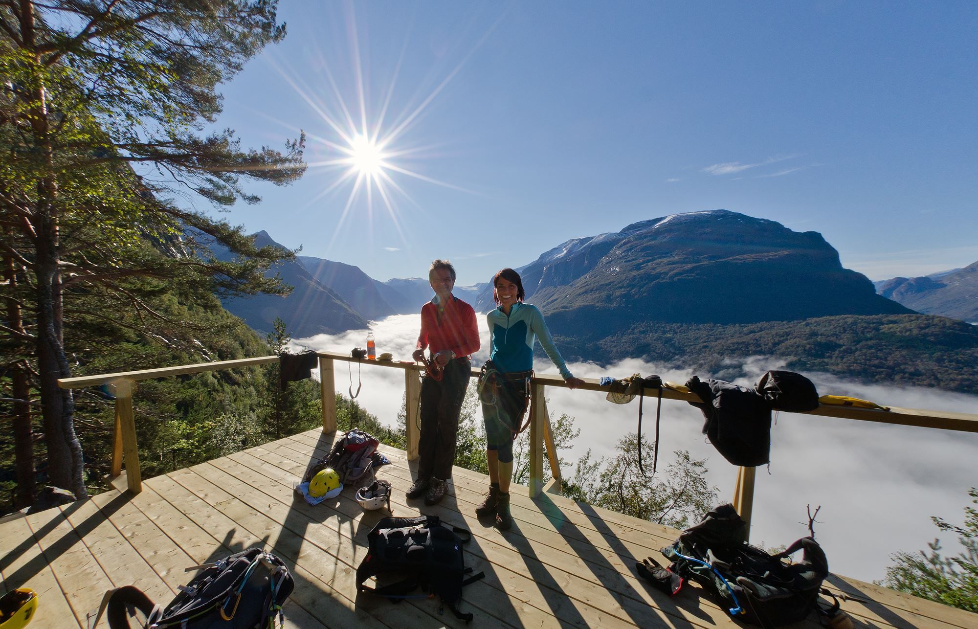 To personar med klatresele står på ei treplatting med tau og utstyr, med sol, fjell og skyer i dalen bak..