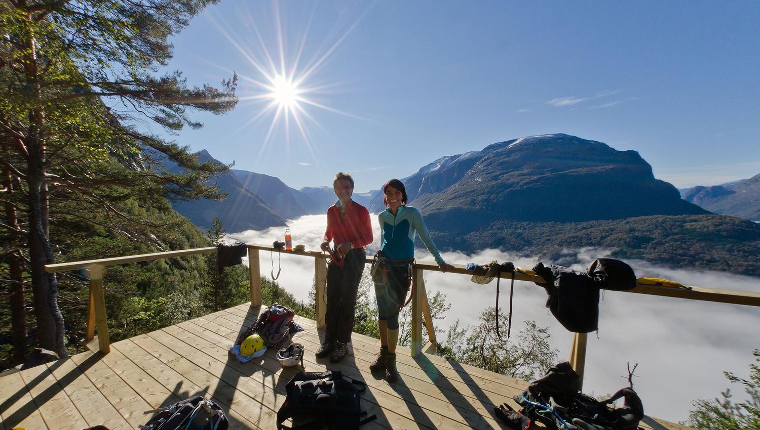 To personar med klatresele står på ei treplatting med tau og utstyr, med sol, fjell og skyer i dalen bak..