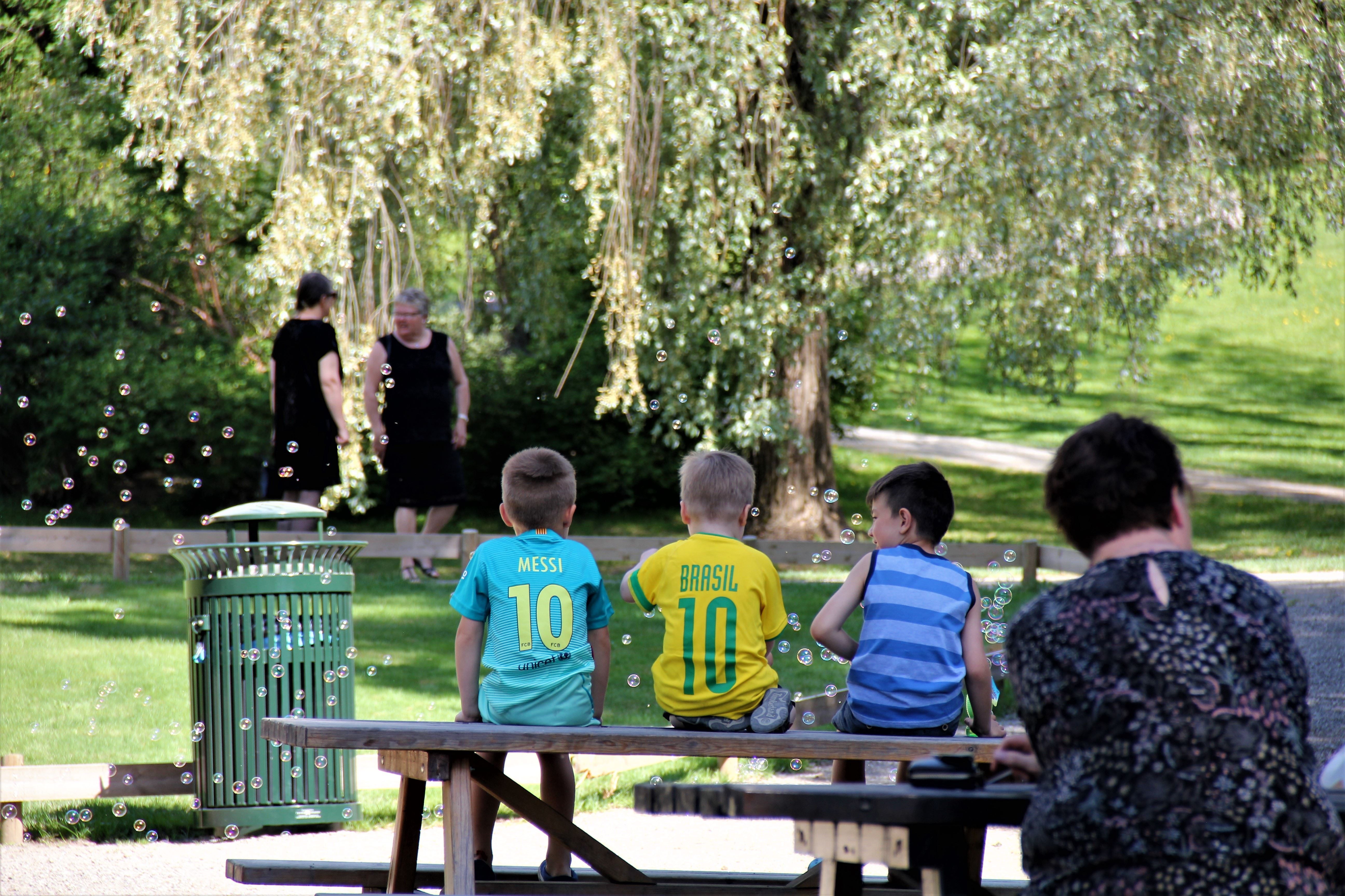 Tre barn sitter på en benk i en grønn park med såpebobler i lufta og trær og stier i bakgrunnen..
