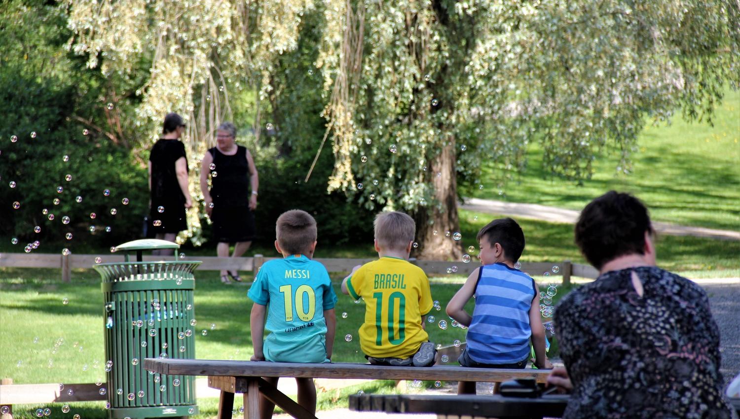 Tre barn sitter på en benk i en grønn park med såpebobler i lufta og trær og stier i bakgrunnen..