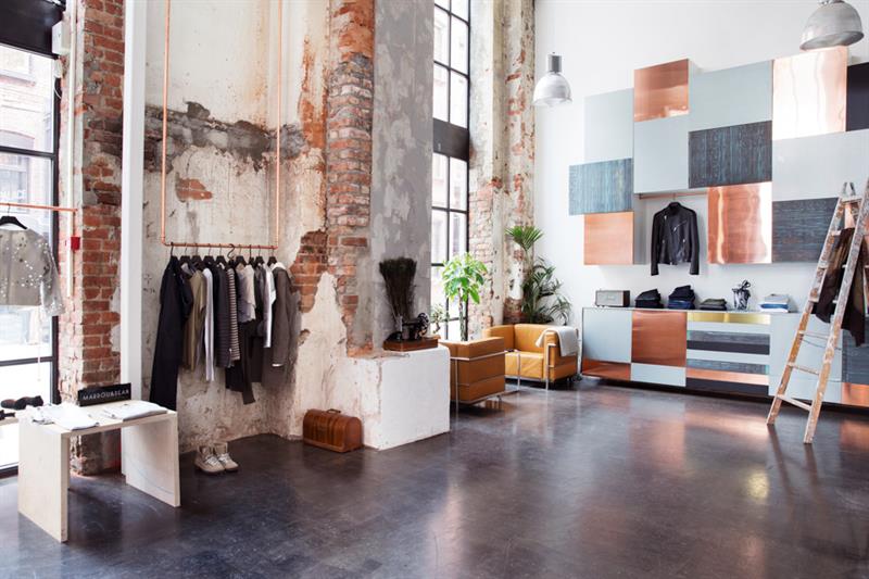 Butikkinteriør med hengende klær, mursteinsvegger, oransje lenestoler, grå taklamper, gardintrapp og veggdekorasjon i grått, sort og bronse.