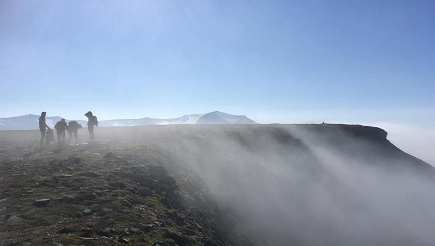 A group of people on top of a mountain with a steep cliff and mist on their right