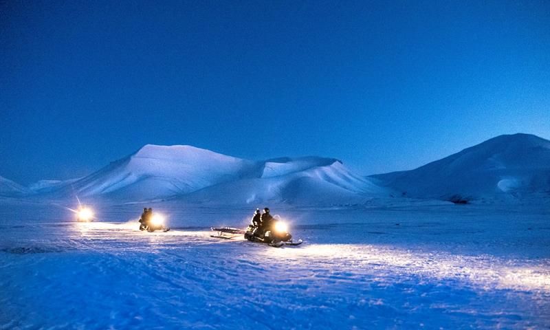 Three snowmobiles driving on the snow. Mountains in the background with dark blue nightsky.