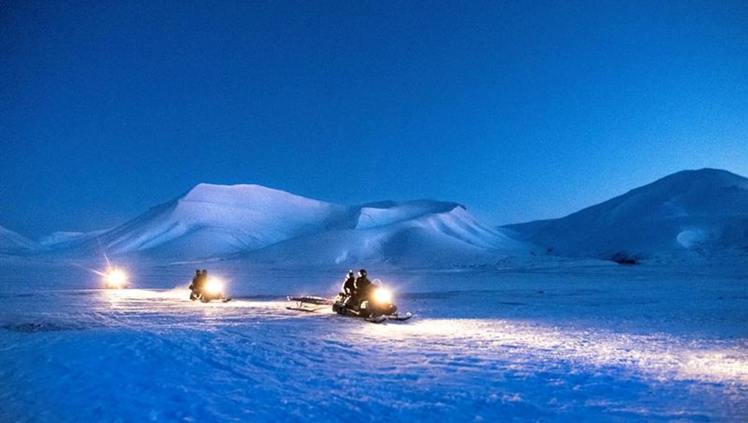 Three snowmobiles driving on the snow. Mountains in the background with dark blue nightsky.