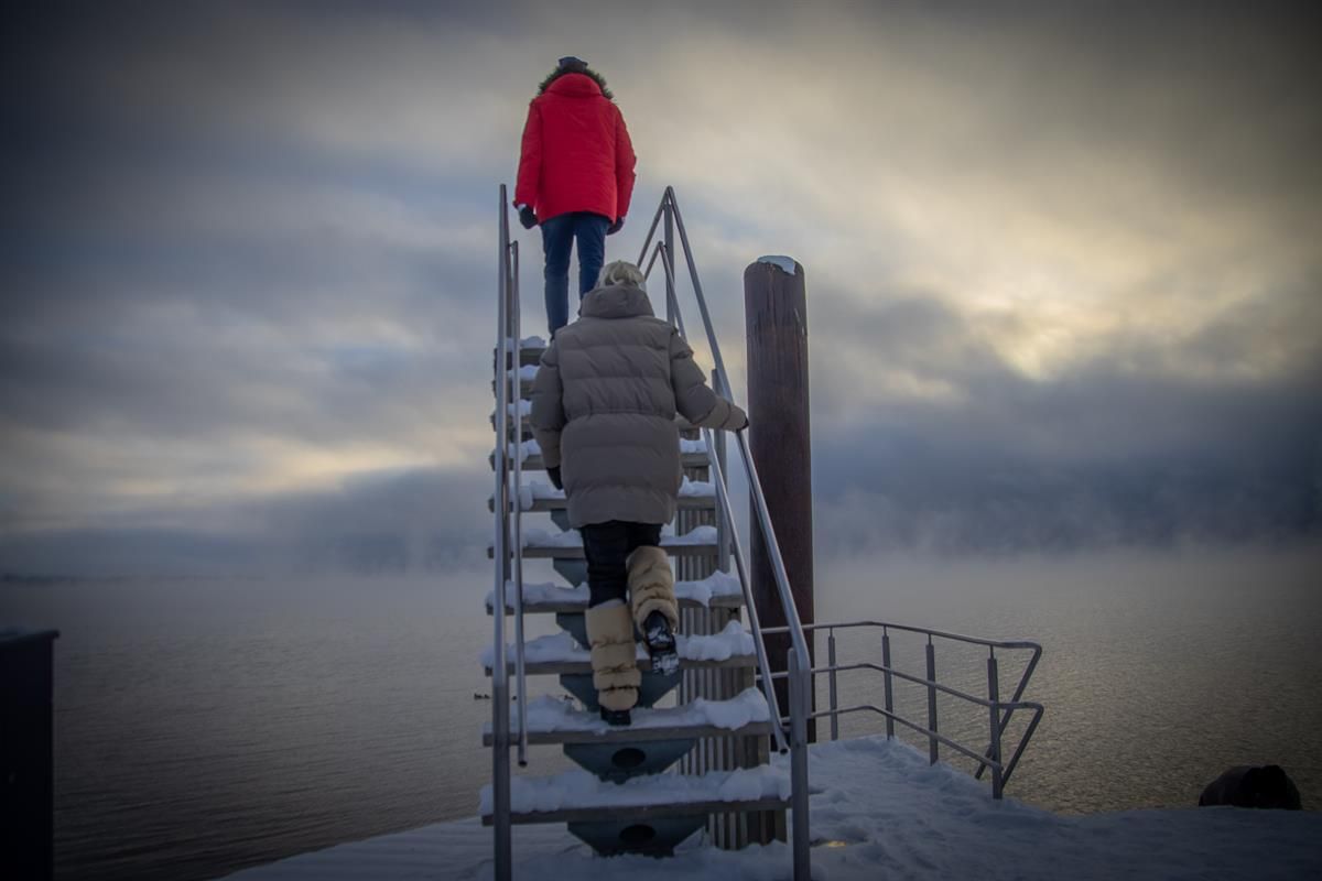 To personer går opp en snødekt metalltrapp ved vannet under en overskyet himmel.