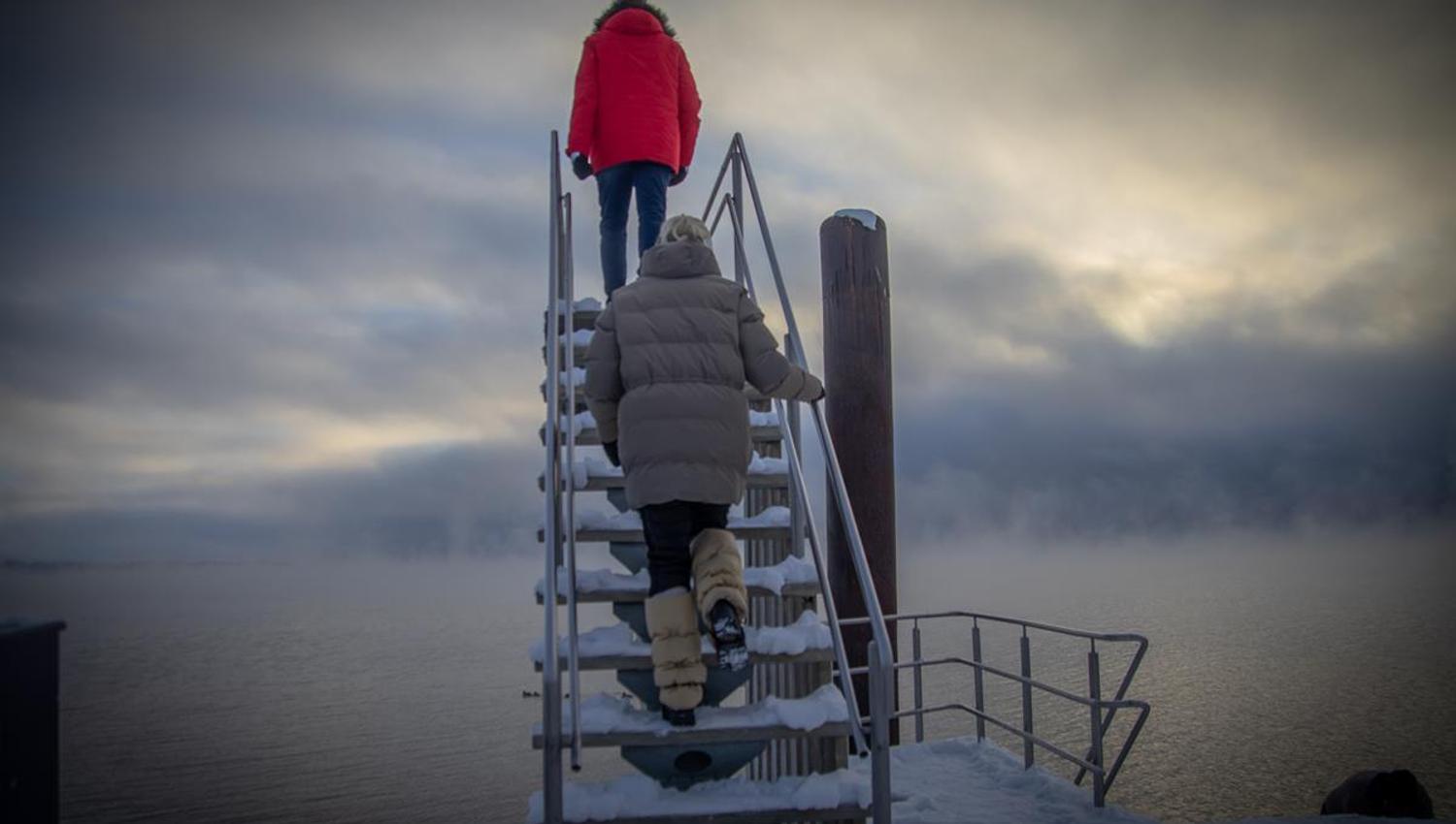 To personer går opp en snødekt metalltrapp ved vannet under en overskyet himmel.