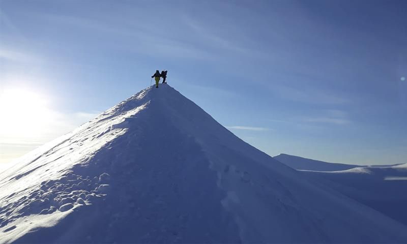 To personer på toppen av en tilsnødd fjellrygg, med snødekte fjelllandskap og en blå himmel i bakgrunnen