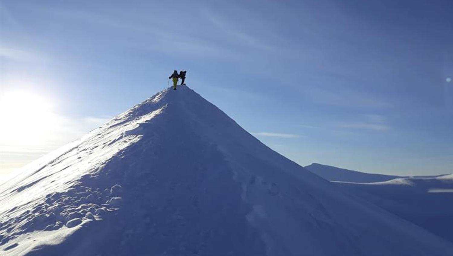 To personer på toppen av en tilsnødd fjellrygg, med snødekte fjelllandskap og en blå himmel i bakgrunnen