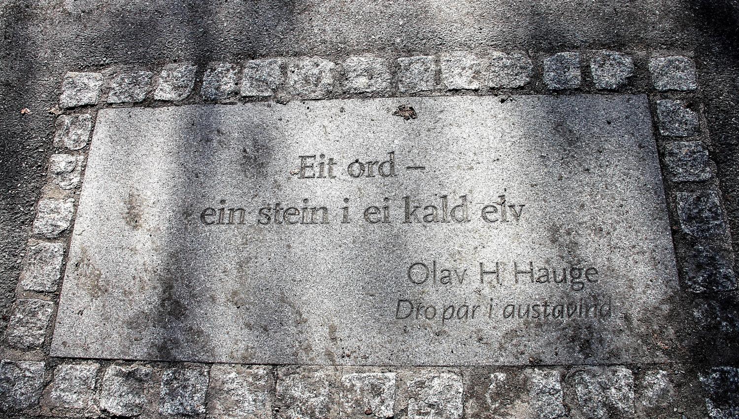 Steinplate i fortau med inngravert tekst, rammet av brostein, med skygger og slitasjemerker på overflaten..
