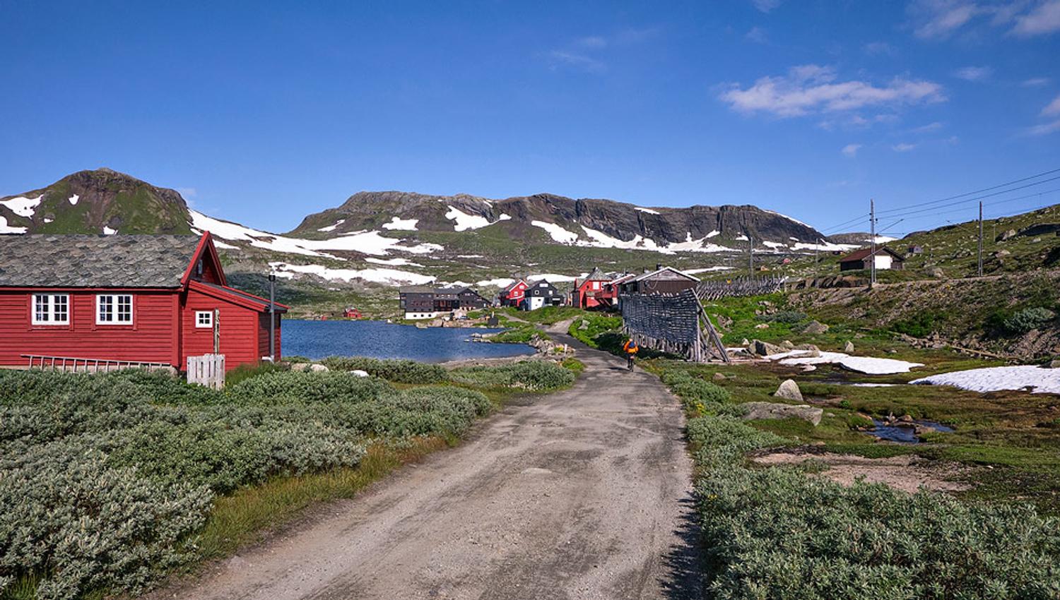 Utsikt mot Finse med raude fjellhytter og Rallarvegen under snødekte fjelltoppar.