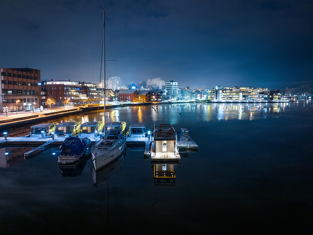 sauna liggene ved elva, med båter på brygge. blokker med lys i bakgrunnen