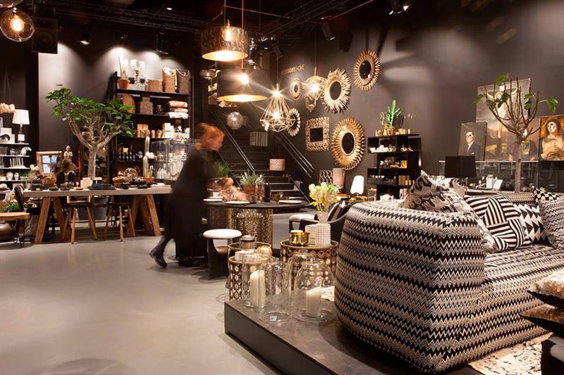 Interiørbutikk med sorte vegger og diverse gyldne lamper, speil, bilder, planter, og en sofa med sort og hvitt mønster.