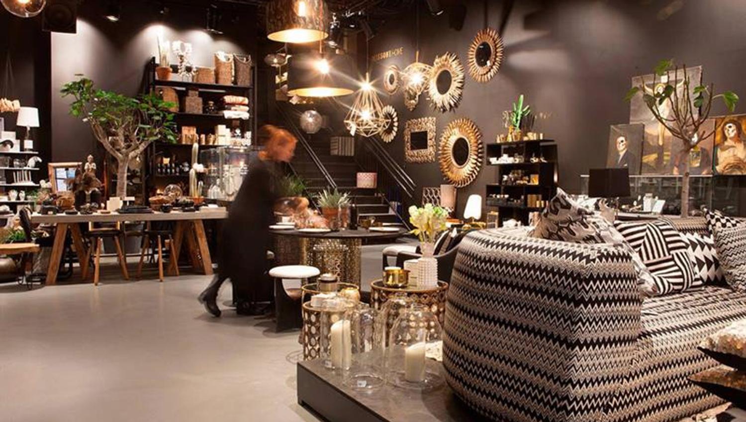 Interiørbutikk med sorte vegger og diverse gyldne lamper, speil, bilder, planter, og en sofa med sort og hvitt mønster.