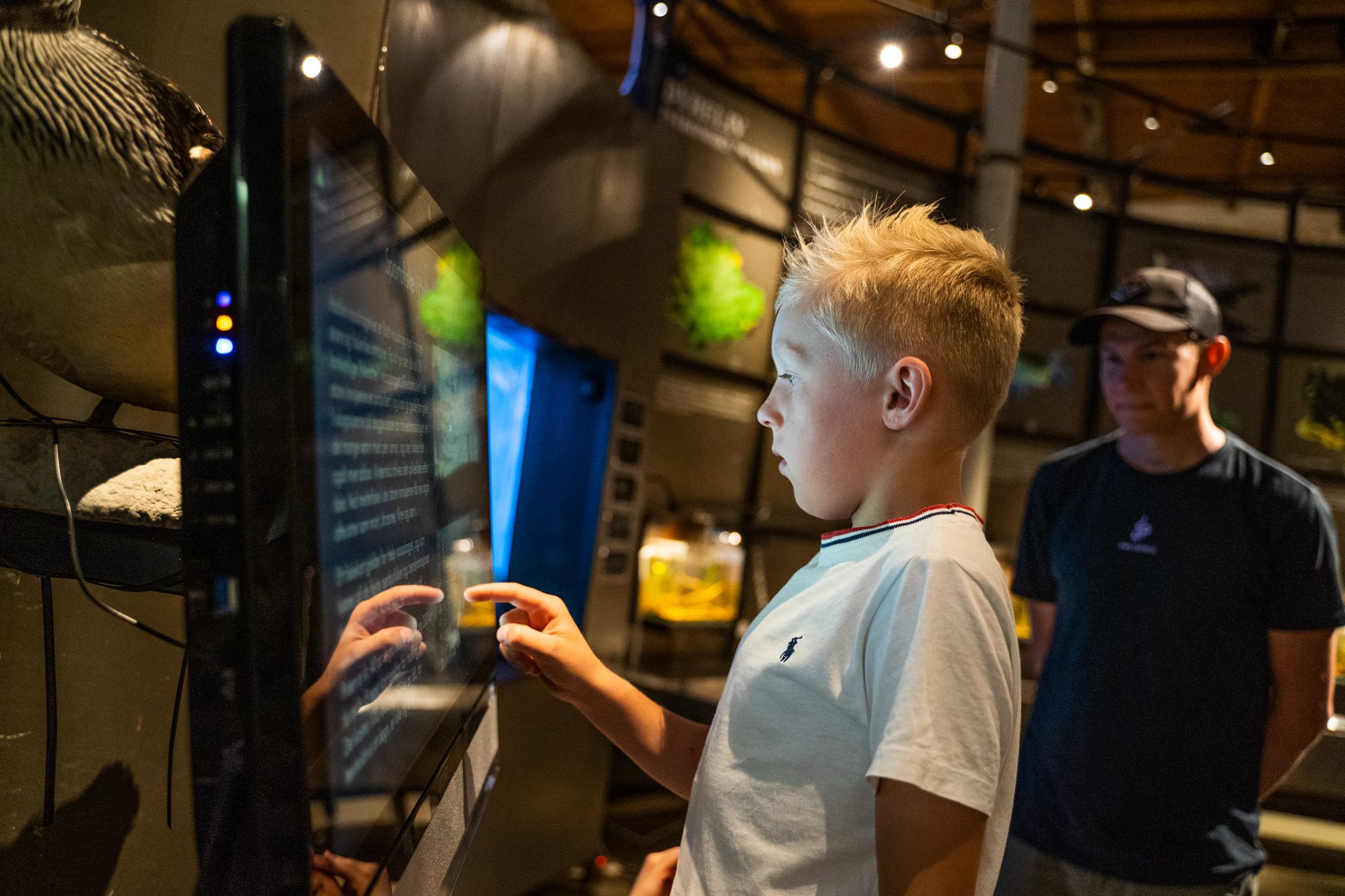 Bildet viser en gutt som trykker på en stor skjerm, kanskje en informasjons- eller læringsskjerm på et museum. Han ser konsentrert ut. Bak ham står en mann og ser på. Rommet er dempet belyst med utstillinger rundt.
