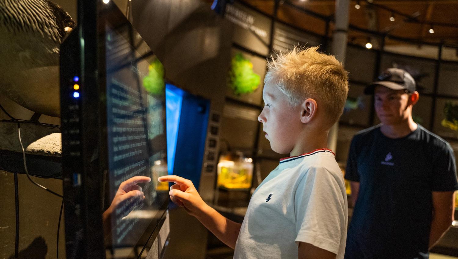 Bildet viser en gutt som trykker på en stor skjerm, kanskje en informasjons- eller læringsskjerm på et museum. Han ser konsentrert ut. Bak ham står en mann og ser på. Rommet er dempet belyst med utstillinger rundt.