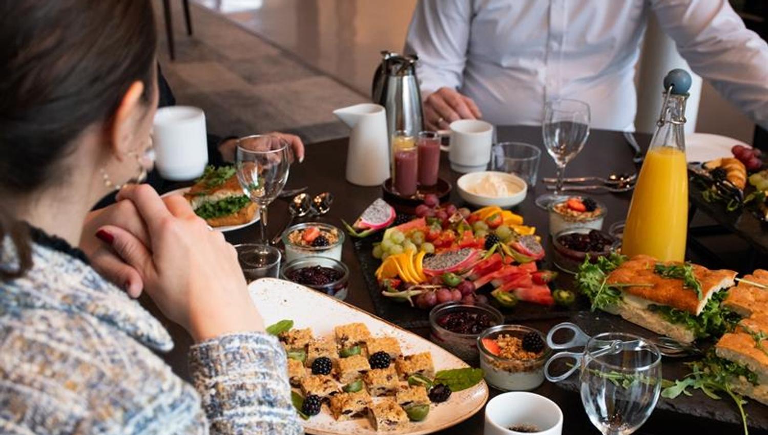 Bord dekket med ulike retter, drikke og kaffe mens flere personer sitter samlet rundt måltidet