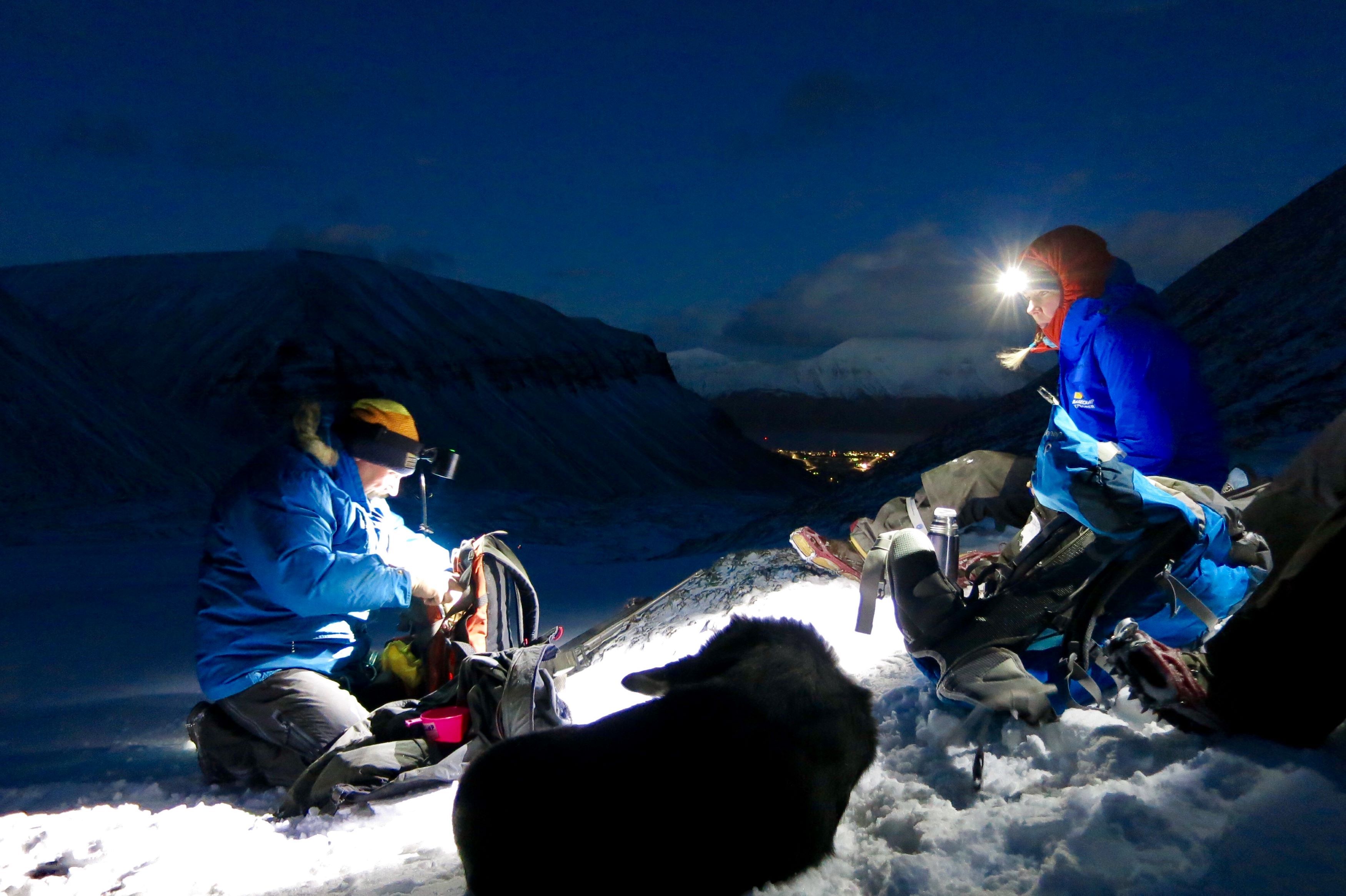 Two people with headlamps and a dog in the foreground, with mountains and Longyearbyen in the background.